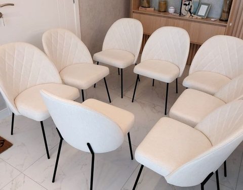 Oito cadeiras de jantar estofadas em tecido off-white, com costura em losango no encosto e pés pretos, reunidas sobre piso de mármore claro